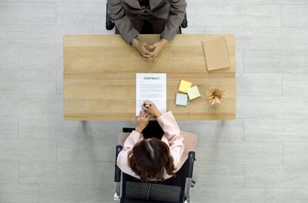 O femeie semnează un document, în fața ei, așezat la masă, se află un bărbat îmbrăcat cu un costul gri.