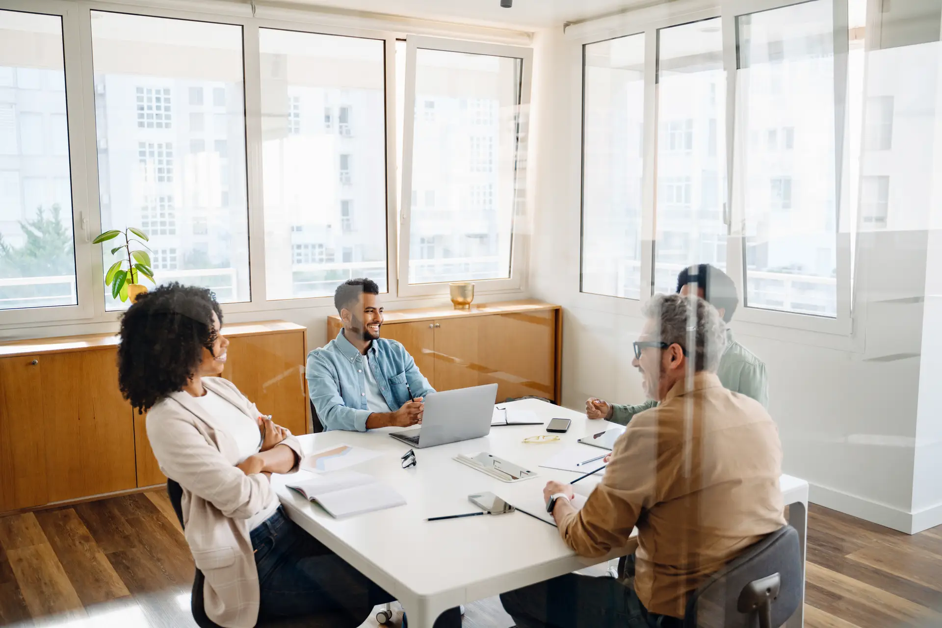A team of four professionals are gathered around a white table in a modern office, indicative of a cooperative and strategic business meeting. View through the glass partition at a diverse work team