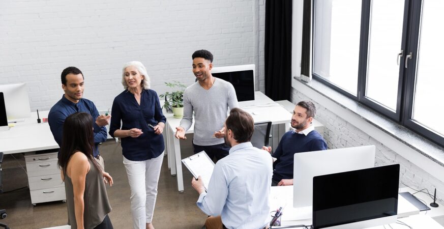 Grup de bărbați și femei discutând într-un birou cu ferestre mari și calculatoare.