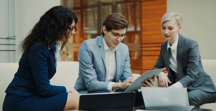 Două femei și un bărbat stau în fața unei măsuțe și se uită pe o tabletă.