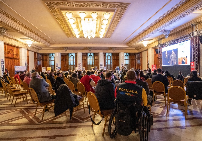 Participanți la un eveniment, printre care și persoane cu dizabilități, vizionează o prezentare pe un ecran.