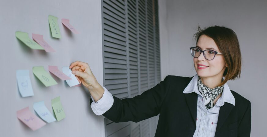 Femeie îmbrăcată elegant lipește notițe colorate pe o tablă de lucru