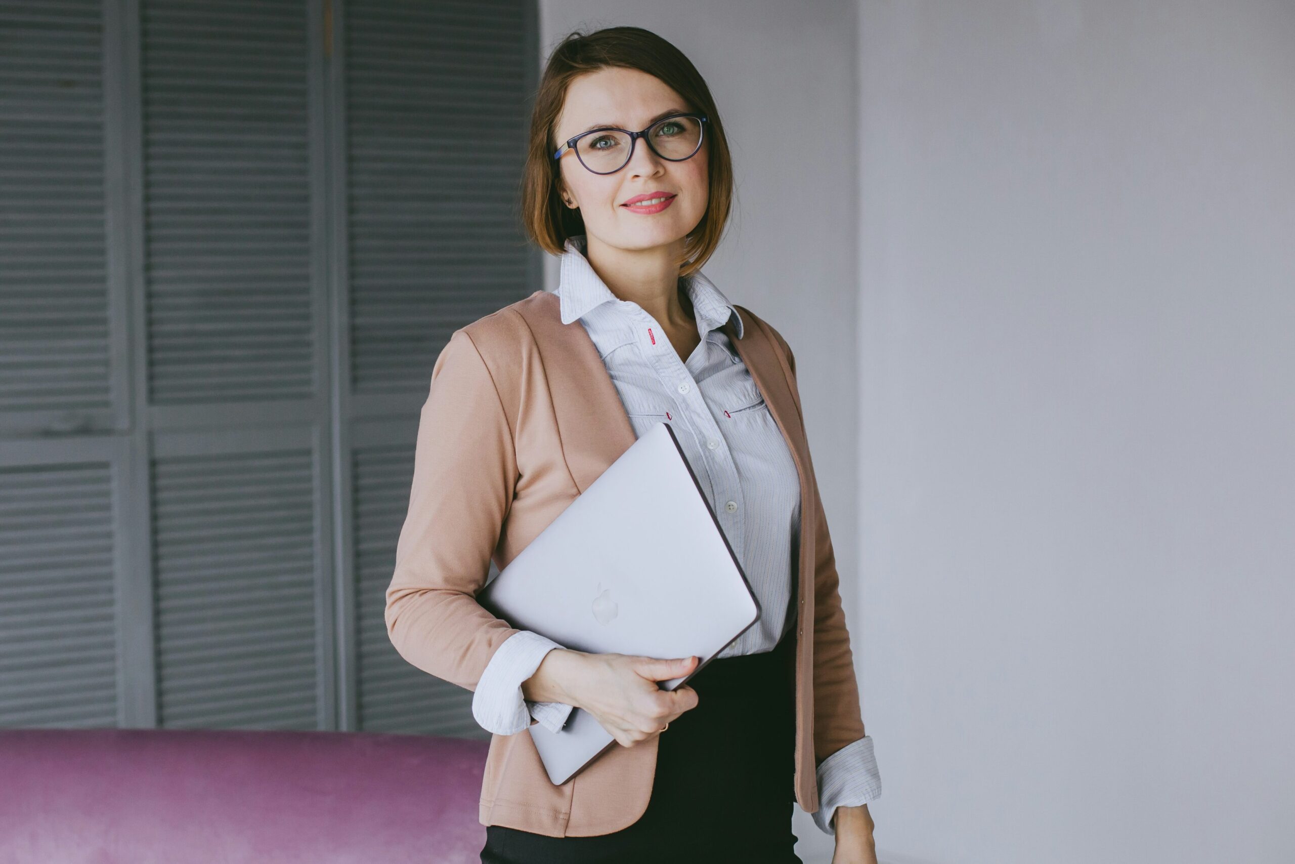 Femeie îmbrăcată elegant ține în mână un laptop și privește drept spre cameră