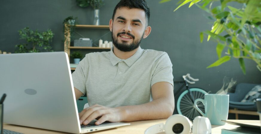Bărbat lucrează la laptop într-un birou luminos, zâmbind spre cameră, cu plante, cafea și căști pe birou.
