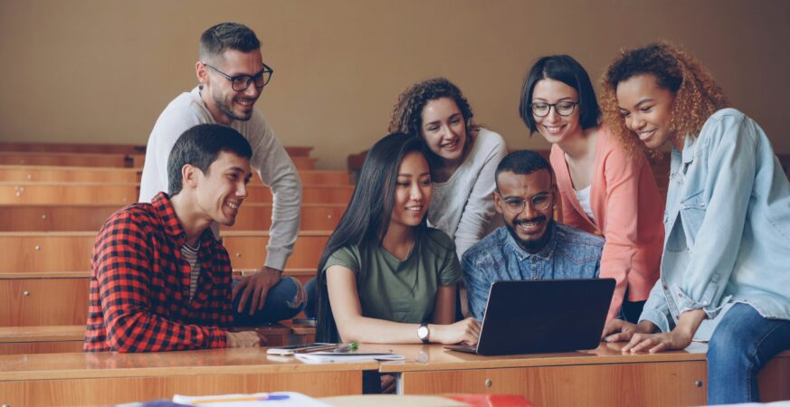 Grup de persoane colaborează într-un amfiteatru, analizând împreună un laptop și zâmbind în timpul lucrului în echipă.