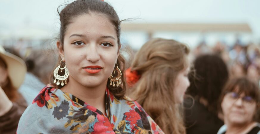 O tânără de etnie romă poartă o cămașă înflorată, cercei mari de aur și o fundă roșie în păr.