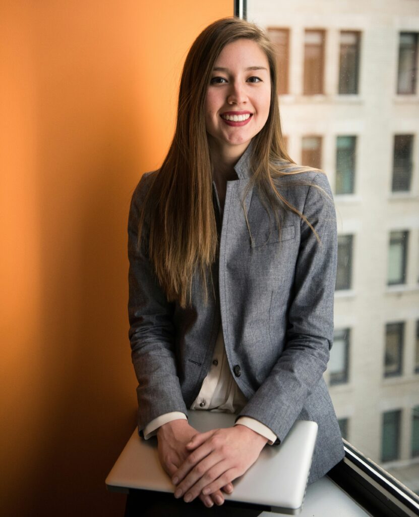 O femeie îmbrăcată cu un costum gri elegant se uită spre cameră și zâmbește, în brațe are un laptop.