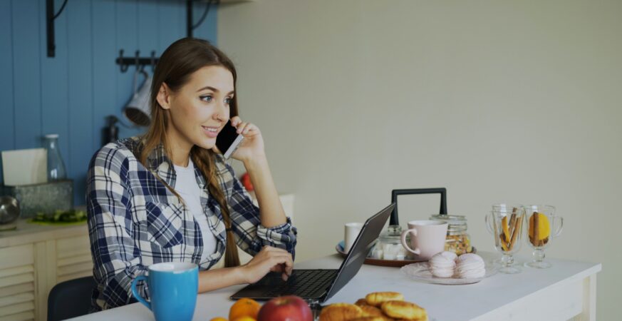 O femeie așezată la masă în bucătărie lucrează la laptop, în timp ce vorbește la telefon.