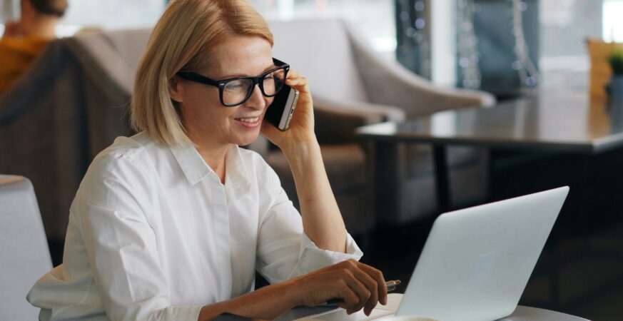 O femeie îmbrăcată cu o cămașă albă elegantă stă la birou și lucrează la laptop, în timp ce vorbește la telefon.